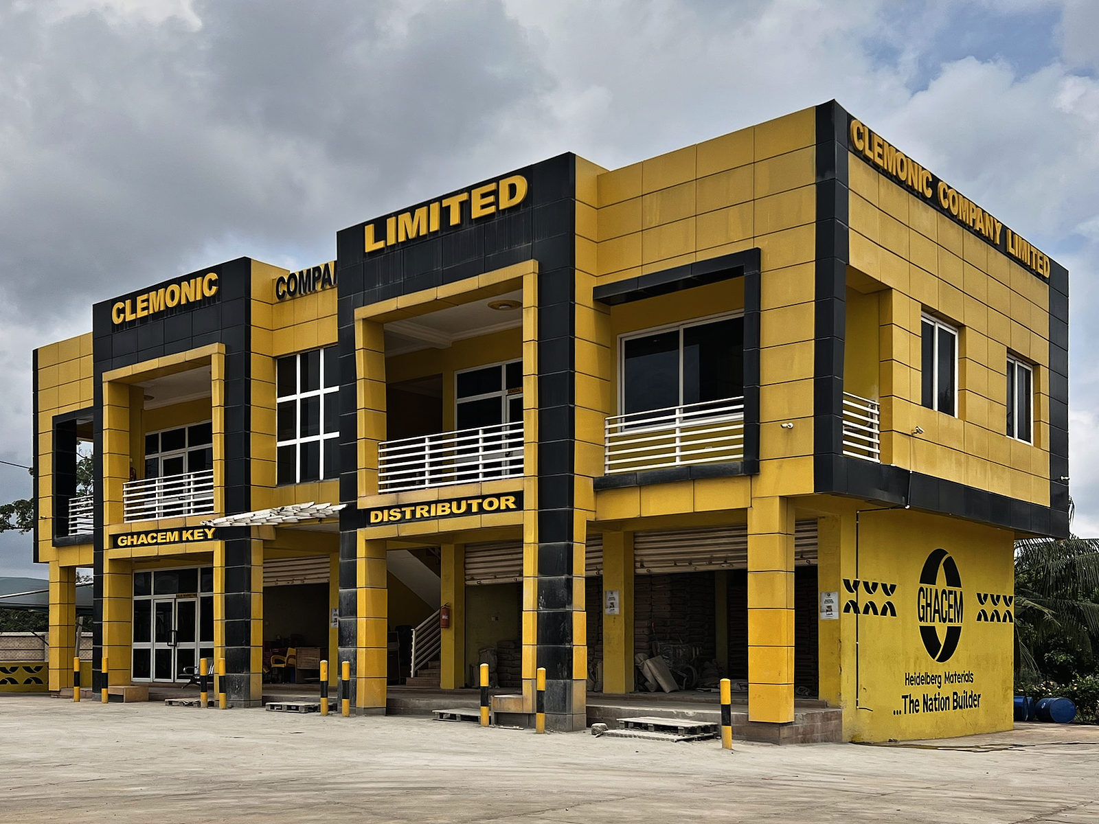Clemonic Company Limited’s two-storey building in Ghacem yellow and black, signed Clemonic Company Limited and Ghacem Key Distributor, with a Ghacem mural on its corner